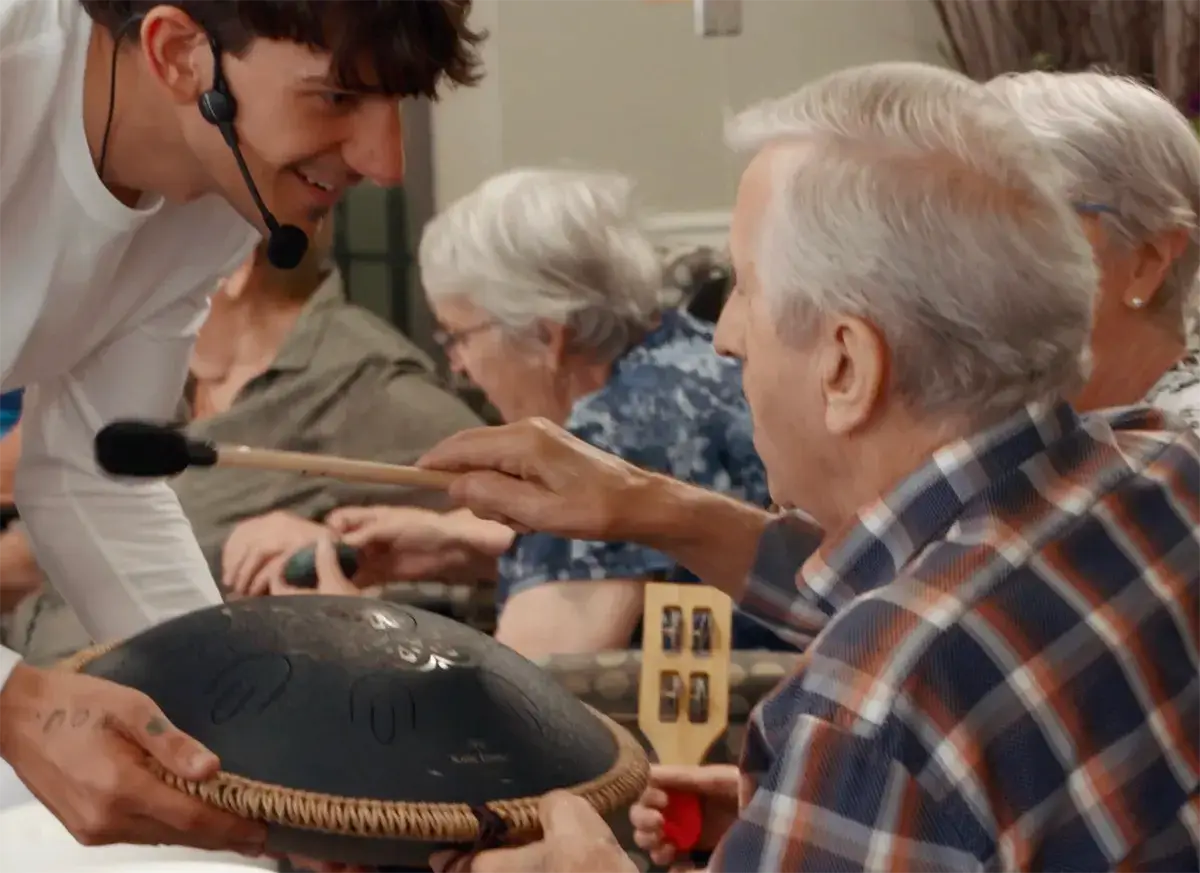 Antoine de Musicofolie avec son handpan, penché vers une participante pendant un atelier en résidence pour aîné·e·s